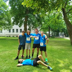 Das Bild zeigt 5 Personen in blauen Tshirts im Park. Vier PErsonen haben die Arme um die Schultern gelegt, die 5. liegt im Gras auf dem Rücken im Vordergrund. Im Hintergrund sind Bäume und das Uniklinikum am Schlossgarten Erlangen zu sehen. .