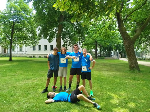 Das Bild zeigt 5 Personen in blauen Tshirts im Park. Vier PErsonen haben die Arme um die Schultern gelegt, die 5. liegt im Gras auf dem Rücken im Vordergrund. Im Hintergrund sind Bäume und das Uniklinikum am Schlossgarten Erlangen zu sehen. .