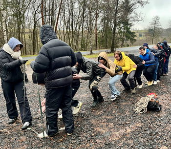 Schulklasse bei einem Teambuilding-Workshop im Freien, Teilnehmer in wetterfester Kleidung, arbeiten zusammen an einer Übung.