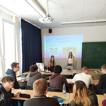 Frau Seischab und Frau Förster vom Finanzamt sprechen vor einer Gruppe Schüler, die interessiert zuhören. Sie zeigen eine Präsentation, im Moment zeigt diese das Wappen von Bayern und Informationen zur Bewerbung beim Finanzamt