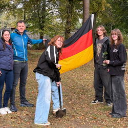Das Bild zeigt die Reisegruppe im Wald. Auf dem Boden liegt etwas Laub. Eine Schülerin ist in der Mitte mit einem Spaten bei der Arbeit, die anderen stehen in einem Halbkreis um sie. Ein Schüler hält eine Deutschlandfahne. Alle schauen in die Kamera