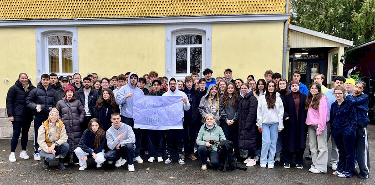 Eine große Gruppe Schülerinnen und Schüler steht vor einem gelben Gebäude. Zwei Schüler in der Mitte halten ein Banner mit dem Text "Studienfahrt Rhön"